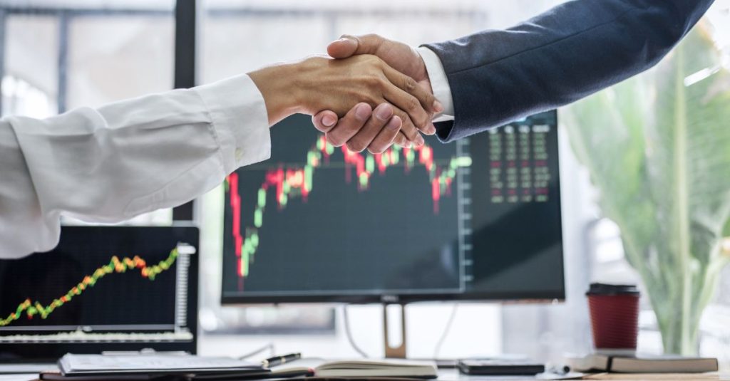 two people shaking hands at the conclusion of a business meeting in front of a computer monitor displaying charts