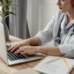 A professional female doctor typing on a laptop in her office with medical notes and stethoscope.