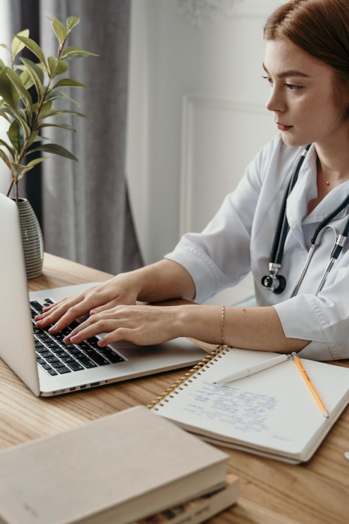 A professional female doctor typing on a laptop in her office with medical notes and stethoscope.